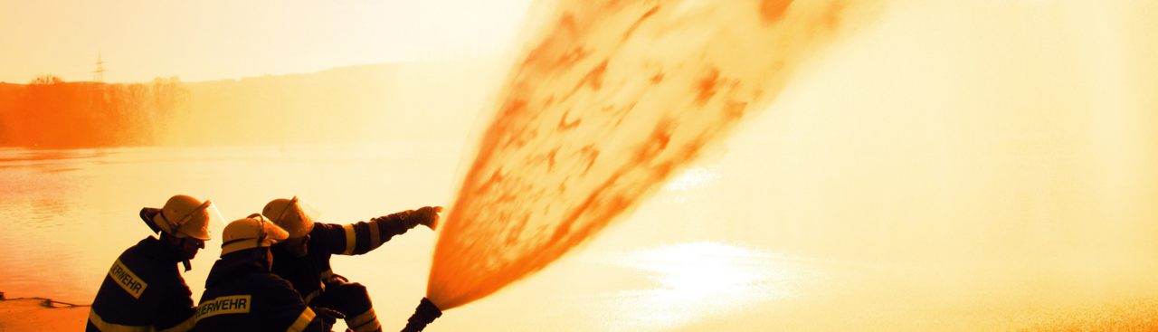 Zwei Feuerwehrangehörige kniend neben einem Wasserwerfer in orange getönt.