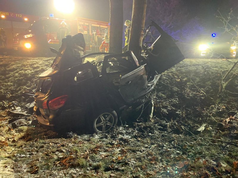 Autowrack um Baum im Vordergrund im Nebel mit Feuerwehr- und Polizeifahrzeugen im Hintergrund