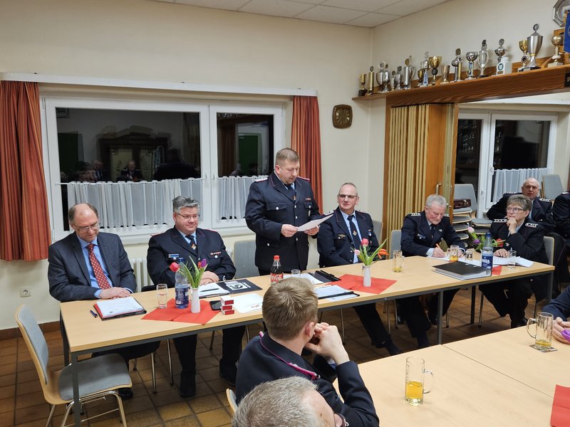 Vorsitzender der Sitzung in der blauen Feuerwehruniform trägt Bericht vor, daneben sitzen die anderen Vorsitzenden der Sitzung