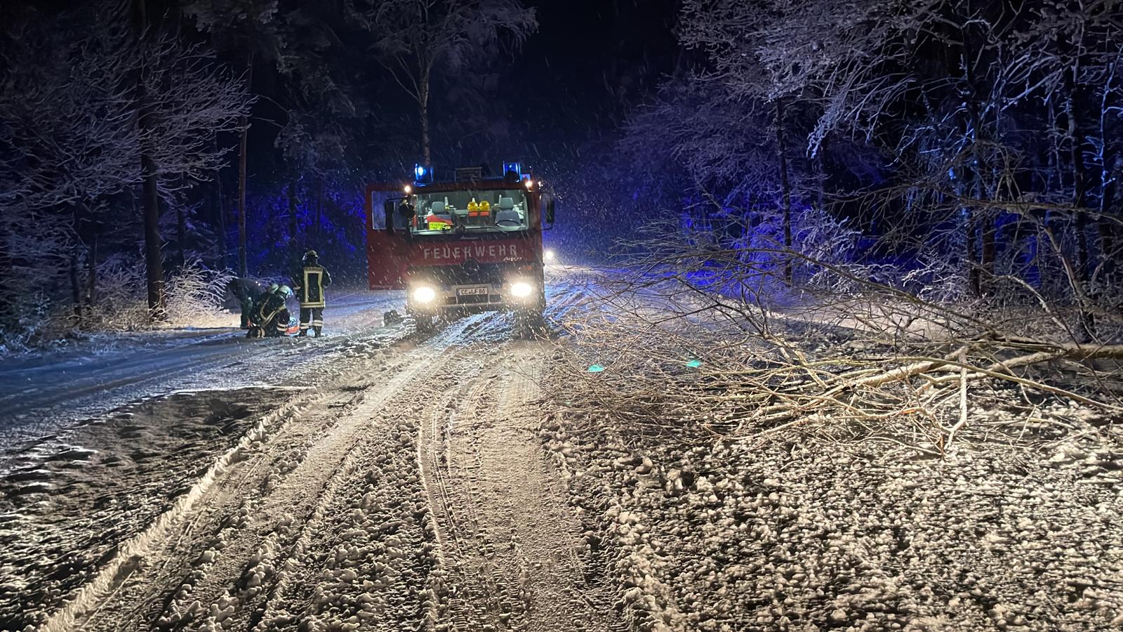 Feuewehrauto auf Straße mit dicker Schneeschicht. Mehrere Feuerwehrangehörige arbeiten links vom Auto bodennah.